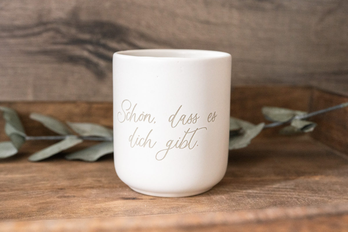 Matte weiße Keramiktasse mit Gravur „Schön, dass es dich gibt.“ in Schreibschrift auf Holzuntergrund, Eukalyptus im Hintergrund.